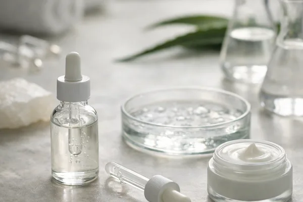 Serumflasche mit Pipette, offener Cremetiegel und Glasschale auf hellem Steintisch, Aloe-Blatt